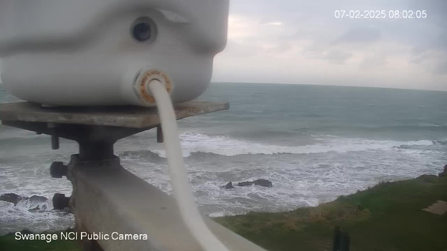 A white container with a hose is positioned on a metal platform, overlooking a turbulent sea with waves crashing against rocky shores. The sky is cloudy, and the scene is set on a coast, with patches of grass visible in the foreground. The time and date are displayed in the upper right corner, indicating early morning.