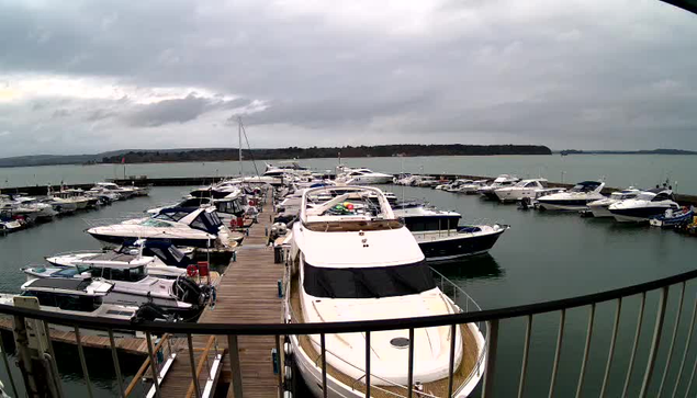 A view of a marina filled with various boats and yachts moored along a wooden dock. The water is calm and reflects the cloudy sky overhead, which suggests overcast weather. In the background, there is a shoreline with trees and possibly hills. The scene conveys a tranquil setting at the harbor.