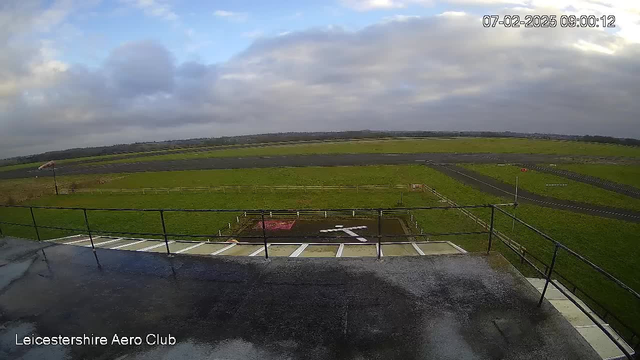 A view from a webcam located at the Leicestershire Aero Club, showing a large open field with a gray runway in the foreground. The runway is bordered by green grass and a few dirt patches. In the lower center, there's a white cross marking on the ground. The sky is overcast with gray clouds, and there is a slight sheen on the surface, indicating recent rain. The timestamp in the corner shows the date and time as 07-02-2025, 09:00:12.