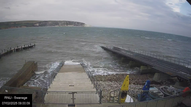 A cloudy day over a coastal scene showing choppy waves in a harbor. There are two jetties extending into the water, one visible to the left and another leading away from the stairs in the foreground. A staircase descends to the water's edge, beside rocky terrain. Kayaks in yellow and blue are secured near the railing. The distant cliffside is visible on the horizon, partially obscured by clouds.