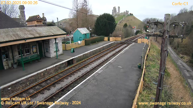 A railway station scene with a platform in the foreground. The platform features a stone building with a sloped roof, and a green bench. Visible is a green information sign and a wooden fence behind the platform. The railway tracks extend into the distance, leading towards a hill with castle ruins atop it. The background contains trees and a small blue shed. The sky is overcast, lending a muted feel to the scene. In the right corner, there is a wooden power pole with wires running along it.