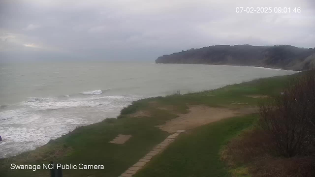 The image shows a cloudy shoreline with the sea on the left side, gentle waves crashing onto the sandy beach. The coastal landscape features green grass and a dirt pathway leading towards the water. In the background, cliffs rise up on the right side, partially obscured by haze. The sky is overcast, indicating a gloomy day. The timestamp in the corner shows the date and time.