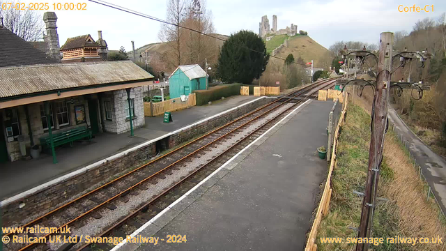 A view of Corfe Castle Railway Station with a platform extending to the right. The platform has a few benches and a sign displaying "WAY OUT." There is a station building with a slate roof and stone walls on the left. In the background, a hill is visible with the ruins of Corfe Castle atop it. Electric poles line the right side of the image, and a narrow road runs alongside the station, bordered by hedges. The sky is mostly clear with a few clouds.