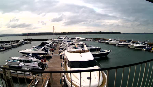 A marina scene showing numerous boats docked in a harbor. In the foreground, several motorboats and yachts are neatly moored along wooden piers. There is a large white yacht at the center with a dark windshield and a deck area. Surrounding boats vary in size and color, with some featuring bright blue and white stripes. The water is calm, reflecting the cloudy sky, and there are distant hills visible on the horizon. The atmosphere appears serene and slightly overcast.