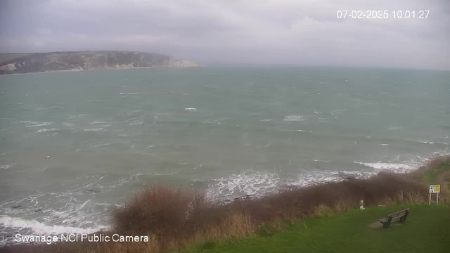 A cloudy coastal scene featuring a turbulent sea with choppy waves. The water appears greenish-blue and is foamy at the edges. In the foreground, there is a grassy area with some bushes, and a bench is visible to the right. A warning sign is placed further along the path. The coastal cliffs are faintly visible in the background, extending into the distance. The overall atmosphere feels windy and unsettled.