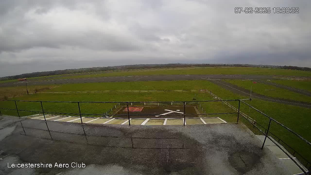 A view from a webcam at Leicestershire Aero Club showing a wide expanse of green grass and a gray, overcast sky. The foreground features a balcony railing, with a helipad marked on the ground below. In the background, there are runways and scattered fencing, with distant hills beyond. The scene appears cloudy and damp, with visible wet areas on the balcony surface.