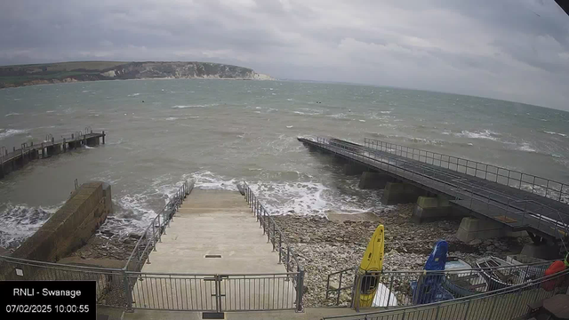 A rocky shoreline with a staircase leading down to the water. Waves are crashing against the rocks. In the foreground, there are two kayaks positioned near the shore, one yellow and one blue. In the distance, a long pier extends into the body of water. The sky is overcast with gray clouds, and a cliff can be seen in the background. The image captures a coastal scene with a sense of movement in the water.