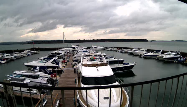 A marina filled with various boats docked in the water. The view shows several white and blue boats moored along wooden docks with some people visible. The sky is overcast with gray clouds, and the water is calm, reflecting the clouds above. In the background, there is a shoreline with trees.