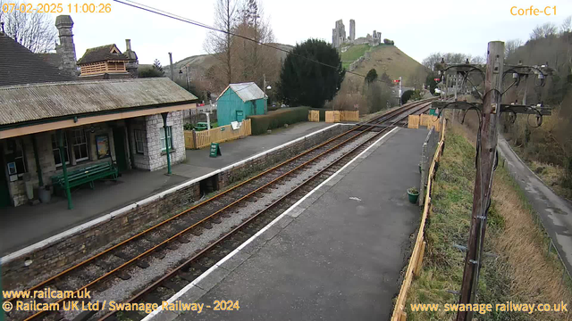 The image shows a railway station platform at Corfe Castle. In the foreground, there are stone walls with a gravel path and railway tracks running parallel to the platform. To the left, there is a stone building with a sloped roof and a green bench. A green shed can be seen behind the building, along with a wooden fence. In the background, Corfe Castle is visible on a hill, surrounded by trees and a clear sky. A wooden telegraph pole is positioned on the right side of the image.