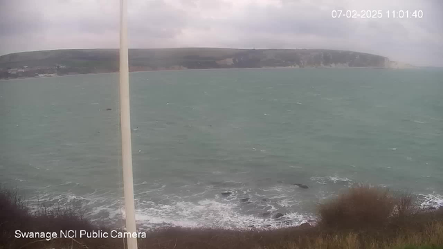 A view of the sea on a cloudy day, with gentle waves lapping against the rocky shore. In the background, green hills rise alongside the coastline. A tall, white post stands on the left side of the image, and there are patches of brown vegetation at the bottom of the frame. The overall atmosphere is tranquil but somewhat overcast. The date and time are displayed in the top right corner.