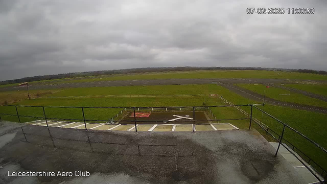 A view from a webcam at Leicestershire Aero Club, showing a large, open green field with a landing strip in the foreground. The sky is overcast with gray clouds. In the lower part of the image, there is a railing with a wet surface, indicating recent rain. There are markings on the ground, including a large white 'X' and a red area nearby, likely indicating an operational zone for aviation activities. The background features a flat landscape extending to the horizon.