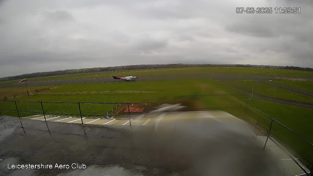 A view from a webcam at Leicestershire Aero Club showing a small aircraft taxiing on a runway. The sky is overcast with gray clouds, and the ground is mostly green grass with some asphalt. In the foreground, there is a railing and wet surface indicating it may have recently rained.