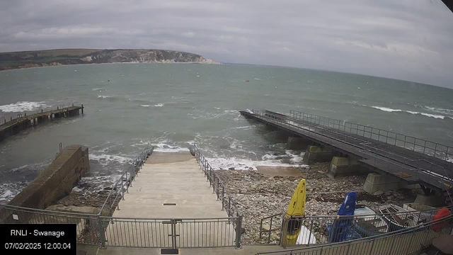 A coastal scene with choppy gray-green water and a cloudy sky. In the foreground, there are stairs leading down to the water, flanked by a fence. To the left, a wooden pier extends into the water. On the right, there are kayaks in blue and yellow colors, and some equipment is stored near the edge. In the background, cliffs rise above the shoreline. The date and time are displayed in the bottom left corner.