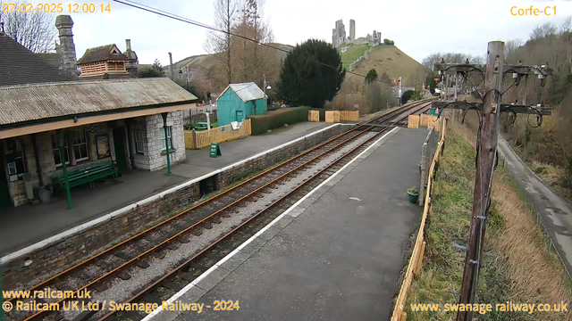 A view of a railway station with a stone platform and a low building featuring a sloped roof. There are several green benches along the platform. In the background, past the tracks, a tall green building and a wooden fence are visible. Beyond that, a hill rises with a castle ruin at its peak, surrounded by trees. A wooden telephone pole stands to the right, with wires and small equipment attached. The sky is cloudy, and the scene is relatively quiet, with no trains in sight.