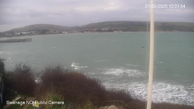 A coastal scene viewed from a webcam, showing a grey, cloudy sky and rough sea. The water is choppy with white-capped waves. In the distance, a small pier extends into the sea, with a few buildings and houses visible along the shoreline. The foreground features some shrubs and vegetation, while the overall atmosphere is calm yet somewhat overcast. The image is timestamped, indicating the date and time.