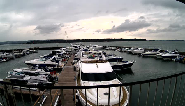 A marina scene featuring several boats docked at a wooden pier. The water is calm and reflects the cloudy sky. In the background, there are rolling hills and trees lining the shore. The light is dim, suggesting an overcast day. Various types of boats are visible, including larger yachts and smaller vessels, all moored in an organized manner.