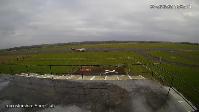 A view from a webcam showing an airfield. In the foreground, there is a railing, and below it, a grassy area with a white "X" marking on the ground, indicating a landing area. In the background, there is a small airplane with a red and black design taxiing on a runway. The sky is overcast with clouds, and the landscape is mostly flat, featuring grass and a distant horizon with tree lines.