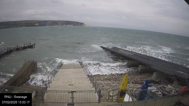 A view of a rough sea taken from a coastal webcam. In the foreground, there are stone steps leading down to the water, flanked by metal railings. To the left, a pier extends into the turbulent water, with several posts visible. On the right side, there are kayaks stored near the water, including yellow and blue ones. The sky is overcast and gray, with waves crashing against the shore and the pier. The image captures the movement and energy of the sea.