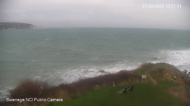 A coastal scene showing turbulent waves crashing against rocky shores under an overcast sky. In the foreground, there is a grassy area with a bench, and a signpost indicating safety rules. The view extends to distant cliffs jutting into the ocean, enhancing the dramatic landscape.