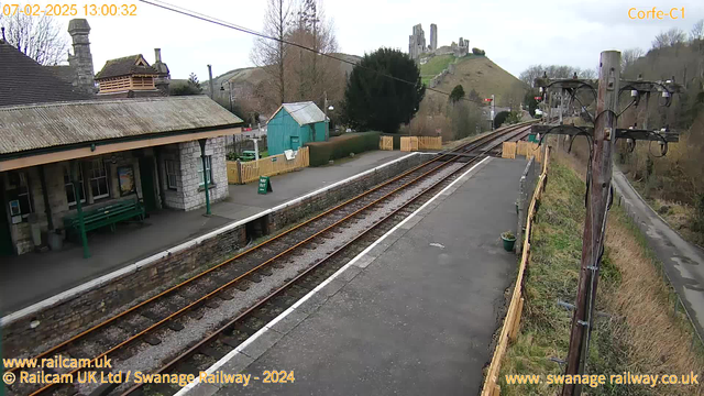 A railway station platform is visible, lined with rustic stone walls and a weathered roof. To the left, there is a green wooden bench and a small building with windows. A wooden fence separates the platform from a grassy area, and there is a green sign indicating "WAY OUT." In the background, a hill features a historical castle or ruin. The railway tracks run parallel to the platform and are lined with gravel, leading towards the horizon. The sky is overcast, contributing to a muted atmosphere. A power pole stands on the right side, with electrical wires visible.