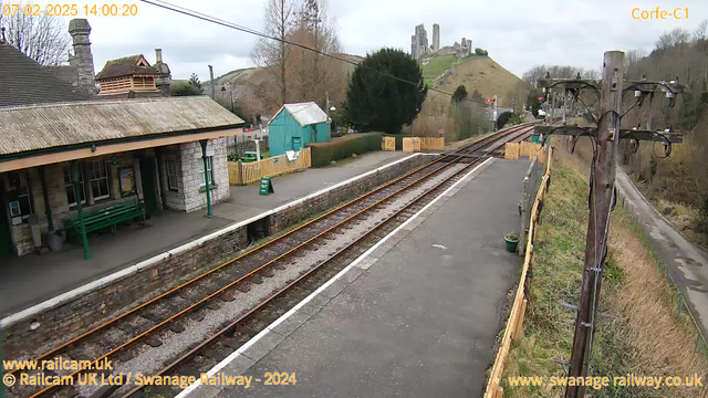 The scene shows a railway station platform with a stone building on the left and a green shed in the background. The platform is empty, with railway tracks running parallel to it. In the distance, a hill is visible, topped by ruins or a castle. There are trees and bushes along the edge of the station area, with wooden fencing partially enclosing the platform. The sky is overcast, suggesting a cloudy day. A sign that reads "WAY OUT" is visible on the platform.