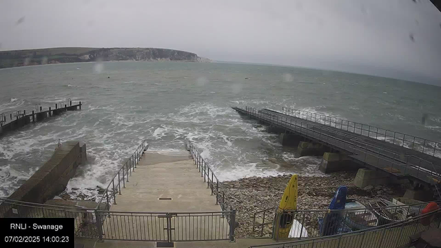 A coastal scene shows turbulent gray waters with waves crashing against a rocky shoreline. In the foreground, there are steps leading down to the water, flanked by metal railings. To the left, a wooden pier extends into the sea, and to the right, a second pier is visible. There are several boats in various colors, including a yellow kayak and a blue one, parked on the shore. The sky is overcast and gray, indicating possibly stormy weather.