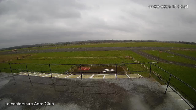 A cloudy sky is visible above a wide, open field with green grass. In the foreground, there is a railing and a solid surface with some wet patches, suggesting recent rain. An airstrip can be seen in the distance, with a helicopter landing pad marked with an "H" symbol on the ground. Some red and white signs are positioned along the edge of the airstrip, and a wooden fence outlines the field area. The scene conveys a tranquil, open-air environment typical of an airfield.