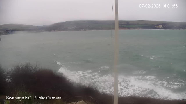 A cloudy, overcast day at a coastal location. The ocean is visible, with waves gently crashing against the shore. In the background, there are rolling hills and a distant cliff. A pole is visible in the foreground, partially obstructing the view. The image is slightly blurred, suggesting mist or rain in the air.