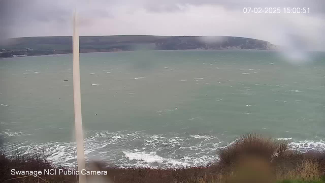 A cloudy sky looms over a choppy sea, with dark green and blue waters. Waves crash against the rocky shoreline, creating white foam. In the foreground, some bushes are visible. The background features a distant cliff and green hills. The image is slightly blurred, suggesting possible rain or mist.