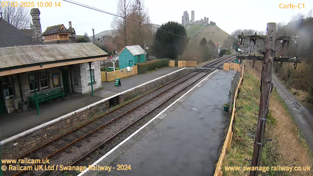 A view of a railway station platform with gray stone walls and a peaked roof. There is a green wooden bench and a sign that reads "WAY OUT." In the background, a dirt path leads into a green area with trees. Electric poles run alongside the railway tracks, which are lined with gravel. In the distance, ruins of a castle can be seen atop a hill, surrounded by green grass and trees. The sky is overcast.