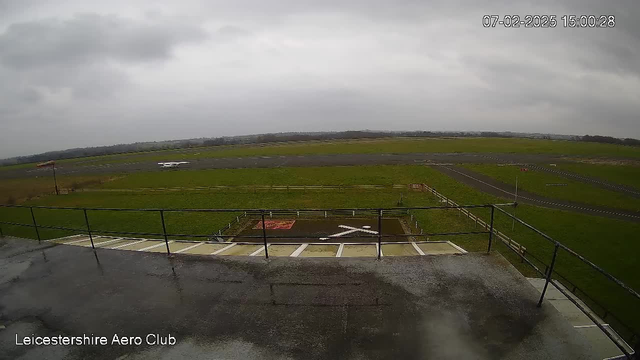 A view from a high vantage point overlooking an airfield. The sky is overcast and gray. In the foreground, there is a railing indicating a balcony or viewing platform. Below, there is a runway with a small aircraft parked to the left on the grass. In the center of the field, there is a large area marked with a red surface, featuring a large white 'X' shape. Surrounding the runway are green fields and a dirt path. The scene appears quiet, with no visible activity.