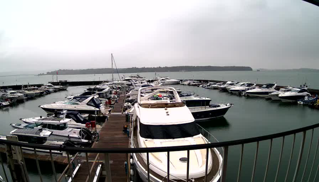 A marina scene featuring numerous boats docked in a calm body of water. The boats are arranged in clusters along wooden docks. The sky is overcast, and the water appears slightly rippled. In the background, a distant shoreline is visible, partially obscured by fog. Several people are seen casually moving about on the docks.