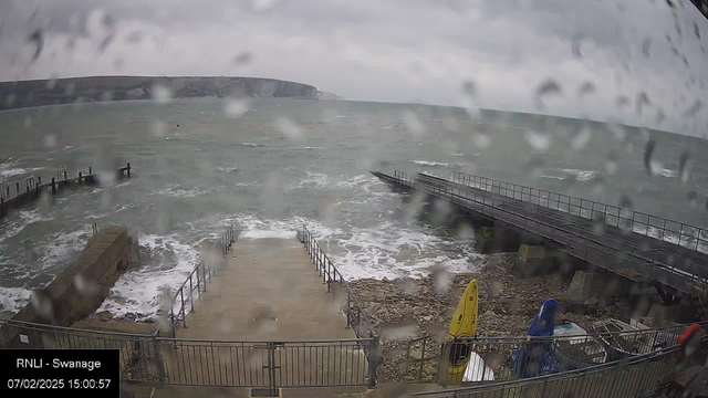 A coastal scene with choppy waves, viewed from a webcam. The image is rainy, with water droplets obscuring the view. In the foreground, there are stairs leading down to the water, flanked by railings. On the left, a short pier extends into the water. To the right, several boats are partially visible, including yellow and blue kayaks. The background features rugged cliffs under a cloudy sky.