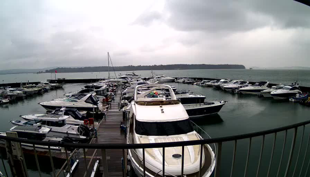 A cloudy scene at a marina with numerous boats docked in the water. In the foreground, there is a large white yacht with a black glass window. Surrounding it are smaller boats of various colors and sizes, primarily in shades of white and blue. The marina is set against a backdrop of grey clouds and a calm sea, with a distant shoreline visible on the horizon. The atmosphere appears overcast and tranquil.