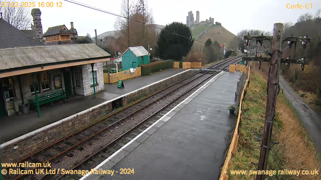 A view of a railway platform at Corfe Castle station, with a stone building featuring a sloped roof on the left. There is a wooden bench and a sign that says "WAY OUT" on the platform. The tracks extend to the right, leading away from the camera. In the background, green hills rise with castle ruins at the top, surrounded by trees. The weather is overcast, and the ground appears wet, suggesting recent rain. There are wooden fences and a small green building on the platform's right side. An electric pole with wires is visible on the far right.