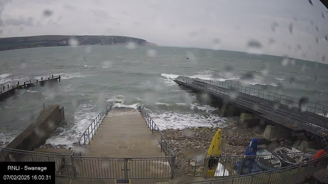 A coastal scene showing rough sea waves crashing near a stone pier. Raindrops can be seen on the camera lens, creating a blurred effect. The pier extends into the water and is lined with metal railings, while a concrete ramp leads down from a higher area to the shore. In the foreground, some fishing or boating equipment in bright colors, including a yellow kayak, is visible. The background features cliffs and hills, partially obscured by cloud cover. The overall atmosphere appears stormy and overcast.