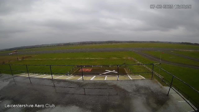 A cloudy sky looms over a wide, grassy airfield. In the foreground, a balcony railing is visible, and below it, a helipad marked with a white 'X' is situated on the ground. To the left, a vehicle is parked beside the airstrip, which runs horizontally across the image. The airstrip is bordered by a wooden fence, and the distant horizon stretches beyond the airfield, creating a serene but overcast scene.