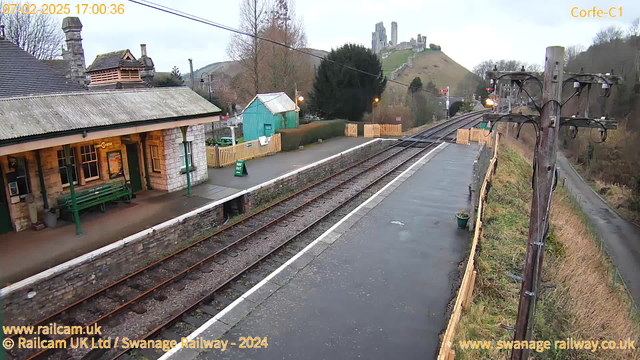 A railway station scene with a stone platform. On the left, there is a building with a peaked roof, large windows, and a sign that reads "Café" above a green bench. A green shed is visible in the background, alongside a wooden fence and a "WAY OUT" sign. The railway tracks extend toward the right of the image, lined with gravel. In the background, a green hill features a historic castle. The sky is overcast, and there are a few trees visible around the station area.