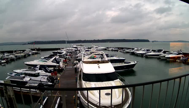 A marina filled with various boats and yachts anchored in calm waters. The sky is overcast with gray clouds, indicating a cloudy day. In the background, a distant shoreline is visible. The marina has wooden docks and several boats are moored closely together, showcasing different sizes and styles.