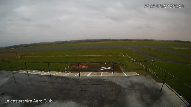 A view from a webcam at the Leicestershire Aero Club shows a large open field with a runway and a grass area. In the foreground, there is a small observation deck with a railing. Below, a red and white striped windsock is visible on the left side, and a large white X marking a helipad is on the ground. The background features a cloudy sky and a distant horizon.