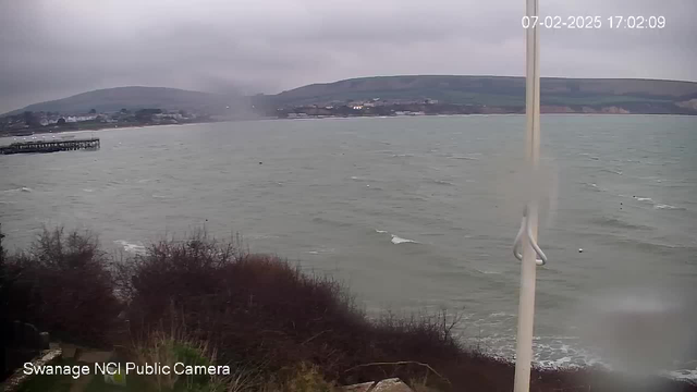 A cloudy day over a coastal scene, with a view of choppy gray waters. In the foreground, there is a small patch of shrubbery. To the left, a wooden pier extends into the water. In the background, gently rolling hills are visible, along with some buildings near the shore. The image appears slightly blurred, possibly due to rain or condensation on the lens. A white pole is positioned on the right side of the image. The timestamp indicates it was captured at 17:02 on February 7, 2025.