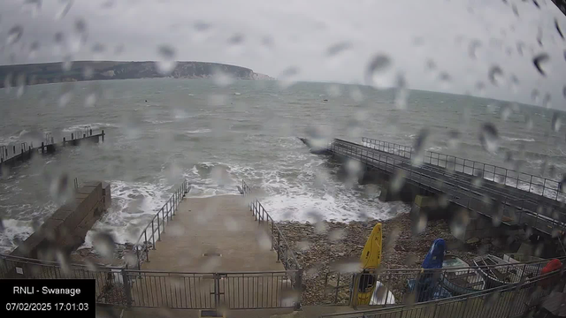 A view of a coastline during a stormy weather, with rain droplets obscuring the lens. The scene shows a pier extending into the choppy sea on the right side, with waves crashing against the rocks and the pier. In the foreground, a concrete ramp leads down to the water, bordered by a metal railing. On the left, there are colorful kayaks (yellow and blue) stored near the shore. The sky is cloudy and gray, indicating a gloomy atmosphere.