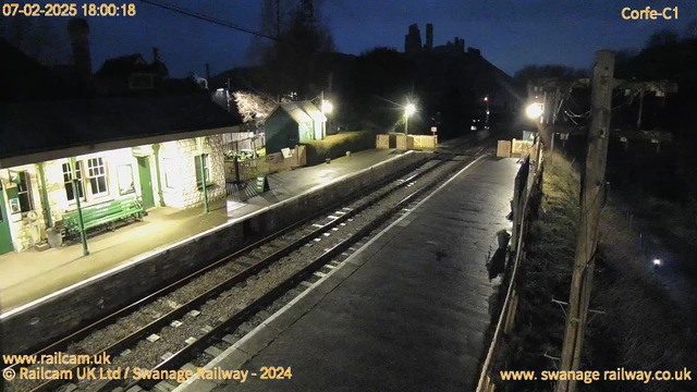 A dimly lit railway station platform at dusk. On the left, there is a stone building with large windows and a green bench outside it. The platform is wet, reflecting the lights and surroundings. Two railway tracks run parallel, leading into the distance. A wooden fence runs along the edge of the platform on the right. There are several light sources illuminating the area, with one particularly bright light near the wooden fence. In the background, silhouetted against the darkening sky, are the outlines of trees and distant hills. The overall atmosphere is quiet and tranquil.