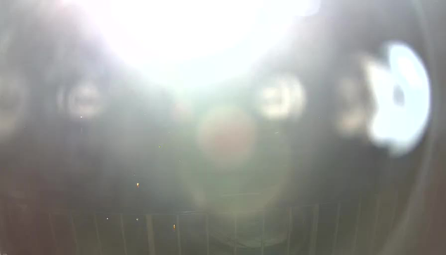 A bright, overexposed image from a webcam showing a blurred view with circular light reflections and faint outlines of objects. There is a prominent glare in the center, making it difficult to discern any detailed features. A horizontal line indicates the presence of a railing or barrier at the bottom of the image.