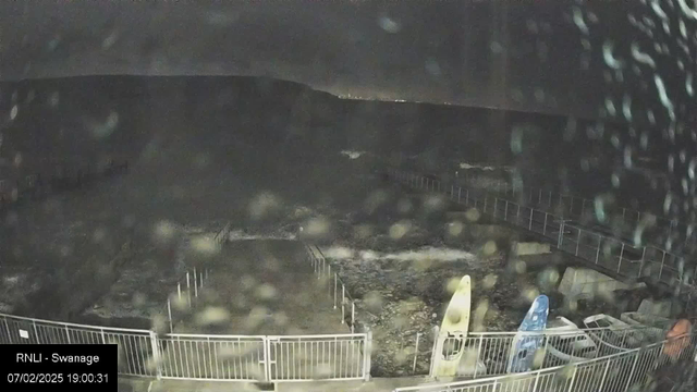 A night view of a seaside location. The image is dark and blurry, possibly due to water droplets on the camera lens. In the foreground, there is a white railing with visible poles and a concrete pathway leading towards the water. Two kayaks, one yellow and one blue, are positioned along the right side of the image. In the background, faint lights from a distant shoreline can be seen against the dark sky. Waves are visible in the water, giving the impression of a turbulent sea.