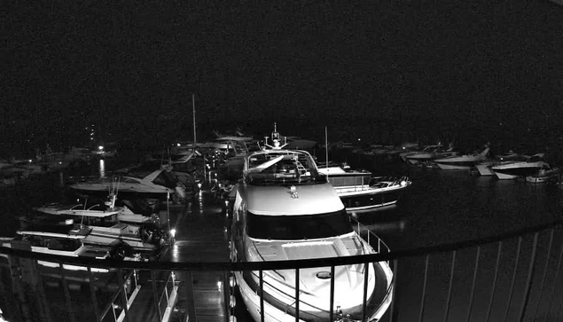 A nighttime view of a marina filled with boats. The scene is predominantly dark, with a few illuminated areas reflecting off the water. In the foreground, a large yacht is docked, facing towards the viewer. Surrounding it are several smaller boats, some partially obscured by shadows. The marina has a wooden walkway that extends towards the water, bordered by a railing. The atmosphere is calm and peaceful, with minimal lighting casting a soft glow on the water and boats.