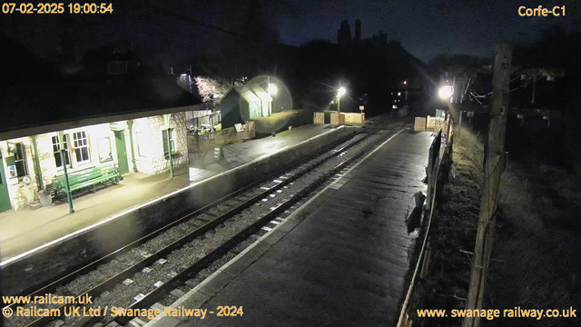 A dimly lit railway station at night features a stone building with large windows and a green bench in front. The platform is wet, reflecting lights from the surroundings. A wooden fence is visible along the right side, with a 'Way Out' sign in front of a small building. The railway tracks run through the center, leading into the darkness beyond the station. A few faint lights illuminate the scene, hinting at the presence of a nearby wooded area and hills in the background.