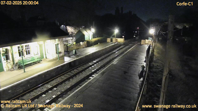 A dimly lit railway station platform at night, with wet pavement reflecting light. On the left, there is a stone building with tall windows and a green bench in front. Opposite the platform, a wooden fence borders the area, with a sign that reads "WAY OUT." The background features trees and a building that appears to be a service shed with green walls. There are limited light sources, including two lamps illuminating the platform and another on the distant right. The scene evokes a quiet, secluded atmosphere typical of nighttime at a rural station.