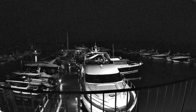 A dimly lit marina at night, featuring several boats docked in the water. Prominent in the foreground is a large boat with a bright white hull and a sleek design. The setting is surrounded by other smaller boats, partially visible amidst the dark water and reflections. The overall ambiance is calm and quiet, with faint outlines of the surrounding structures.