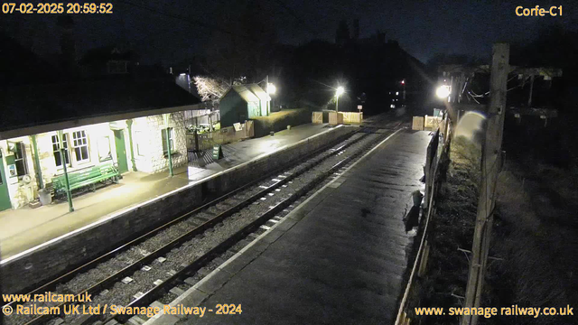 A dimly lit railway station at night. The platform is mostly empty and wet, reflecting light. On the left side, there is a stone building with large windows and a green bench outside. A railway track runs along the bottom of the image with a signal light visible in the distance. There is a fence with a sign that says "WAY OUT" near the right edge of the platform. The setting appears quiet with some faintly lit trees in the background.